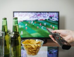 Person sitting at table with beer and chips watching Super Bowl holding remote and point it at the TV